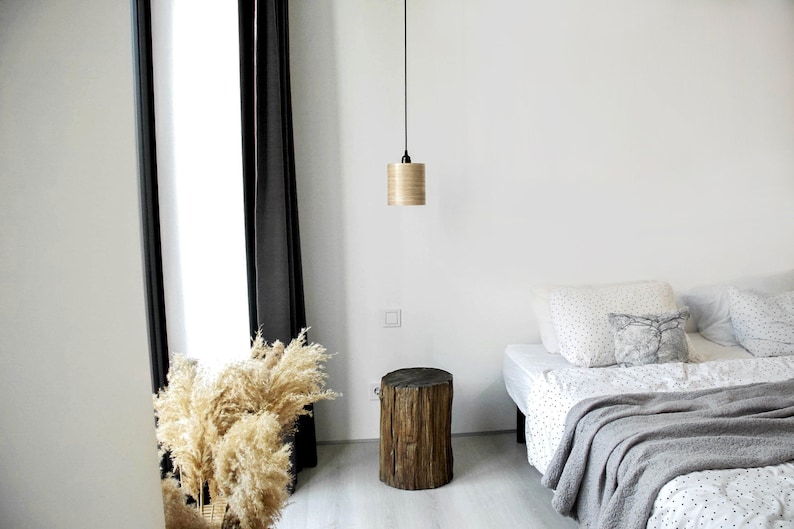 May include: A bedroom with a white wall, a window with black curtains, a wooden side table, a bed with a white and black patterned duvet, a gray blanket, and a light fixture with a wooden shade.