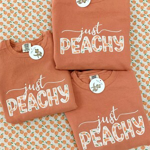 May include: Three peach-colored sweatshirts with the phrase "just PEACHY" printed on the front in white script and peach-patterned letters. Each sweatshirt has a round tag attached. The background is a white fabric with a peach pattern.