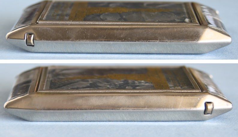 Op de afbeelding: Een rechthoekige zilverkleurige metalen doos met een scharnierend deksel. Het deksel heeft een gedetailleerd, mogelijk gegraveerd ontwerp met grijstinten en goud. De doos heeft een gepolijste afwerking en een veilige sluiting.