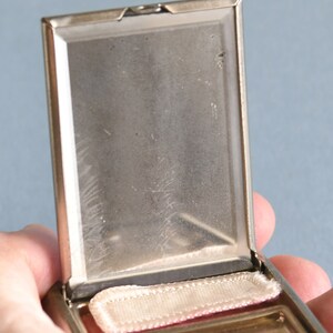 Op de afbeelding: Open, vintage zilverkleurige compact met een rechthoekige vorm. De binnenkant heeft een spiegel en een kleine, rechthoekige poederdons. De compact is opengehouden, waardoor de details van het interieur zichtbaar zijn. De achtergrond is effen lichtblauw.