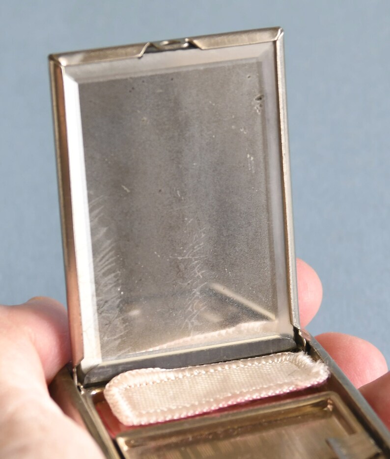 Op de afbeelding: Open, vintage zilverkleurige compact met een rechthoekige vorm. De binnenkant heeft een spiegel en een kleine, rechthoekige poederdons. De compact is opengehouden, waardoor de details van het interieur zichtbaar zijn. De achtergrond is effen lichtblauw.