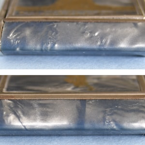 Op de afbeelding: Close-up van een rechthoekige, zilverkleurige metalen koffer met een blauwgrijze, getextureerde oppervlakte. De koffer heeft een goudkleurige rand rond een centraal paneel. De hoeken zijn afgerond en de randen zijn licht gebogen.