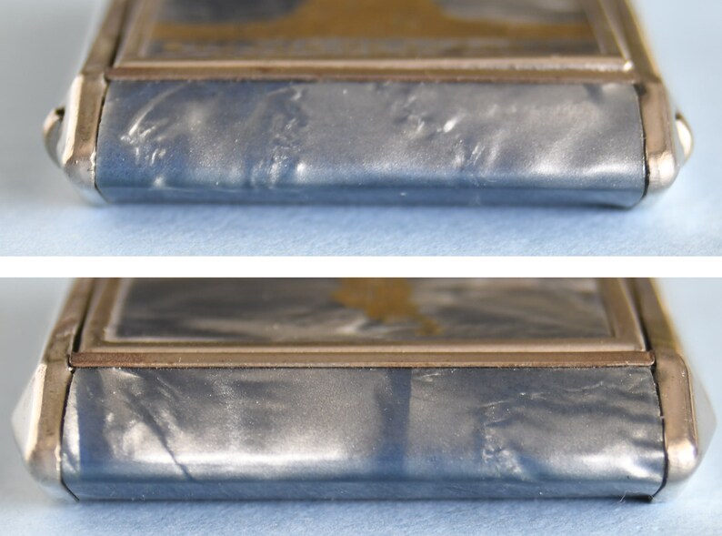 Op de afbeelding: Close-up van een rechthoekige, zilverkleurige metalen koffer met een blauwgrijze, getextureerde oppervlakte. De koffer heeft een goudkleurige rand rond een centraal paneel. De hoeken zijn afgerond en de randen zijn licht gebogen.