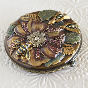 Op de afbeelding: Een ronde, decoratieve compact met een bloemenontwerp. De compact heeft een grote, gedetailleerde bloem met bruine en paarse bloemblaadjes, geaccentueerd met roze en heldere kristallen. Een bij zit op de bloem en groene bladeren omringen de bloem.