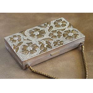 May include: A rectangular silver-toned clutch with a floral design. The top of the clutch features an embossed pattern of flowers and leaves with a beaded texture. A gold-colored chain is attached to the side.