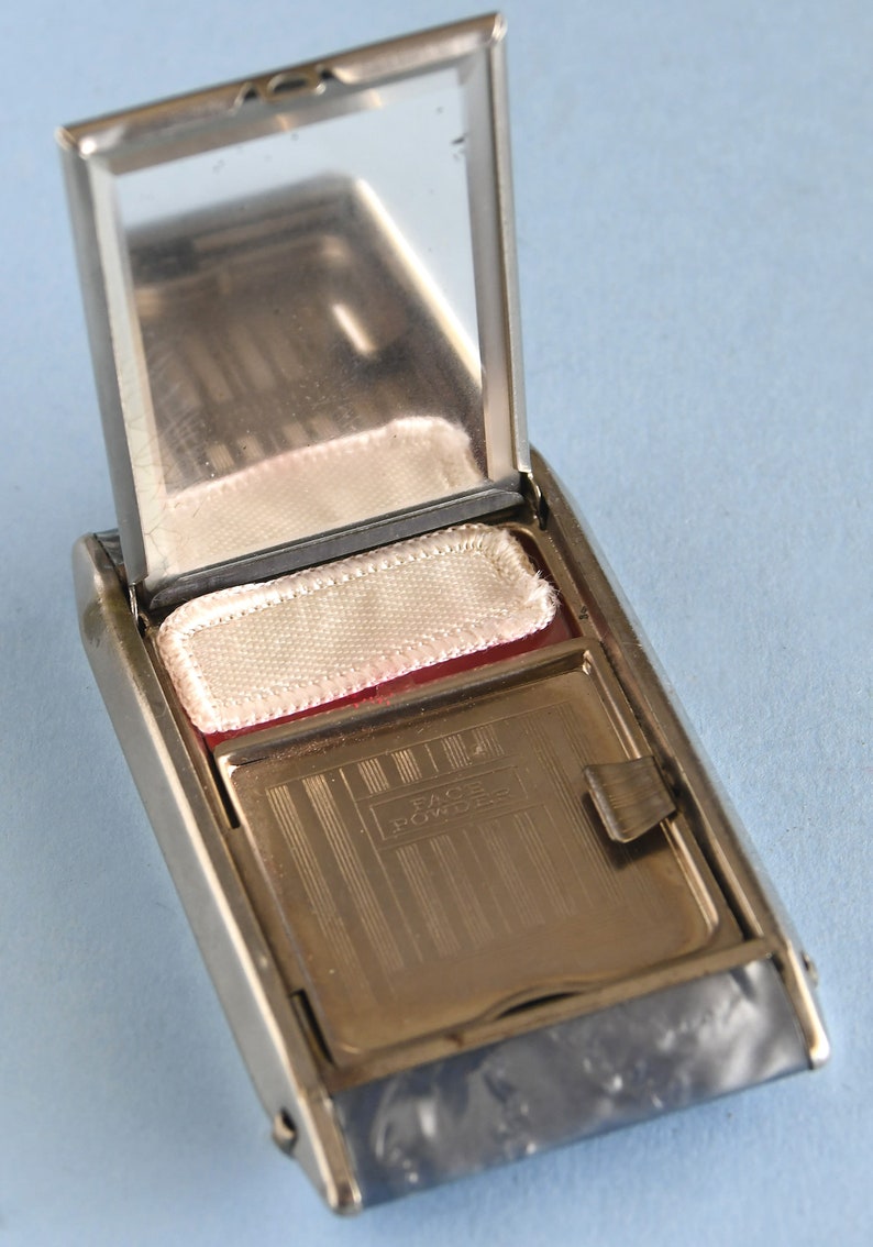 Op de afbeelding: Open zilverkleurige compactpoederdoos met een spiegel, poederdons en een compartiment met het opschrift "FACE POWDER". De rechthoekige compactdoos heeft afgeronde randen en een scharnierend deksel. De poederdons is wit. De achtergrond is lichtblauw.
