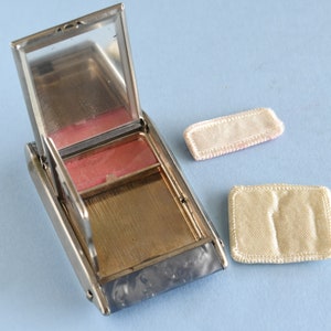 Op de afbeelding: Een open, rechthoekige, zilverkleurige compactpoederdoos met een spiegel en een roze blushcompartiment. Twee kleine, rechthoekige, cr&egrave;mekleurige poederdonsjes liggen ernaast. De compactdoos ligt op een lichtblauwe ondergrond.