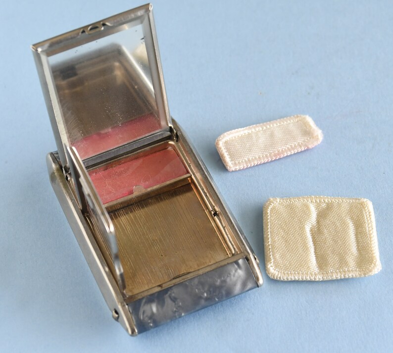 Op de afbeelding: Een open, rechthoekige, zilverkleurige compactpoederdoos met een spiegel en een roze blushcompartiment. Twee kleine, rechthoekige, cr&egrave;mekleurige poederdonsjes liggen ernaast. De compactdoos ligt op een lichtblauwe ondergrond.