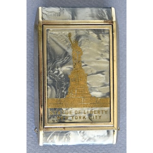Op de afbeelding: Een rechthoekige, zilverkleurige sigarettenetui met een goudkleurig frame. De voorkant heeft een goudkleurige afbeelding van het Vrijheidsbeeld met de tekst "STATUE OF LIBERTY NEW YORK CITY". De etui heeft een parelmoer, gemarmerd ontwerp.