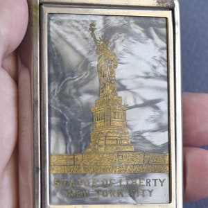Op de afbeelding: Een rechthoekige, zilverkleurige metalen behuizing met een goudkleurig ontwerp van het Vrijheidsbeeld. De achtergrond heeft een gemarmerd, parelmoerachtig effect. De tekst "STATUE OF LIBERTY NEW YORK CITY" staat eronder.