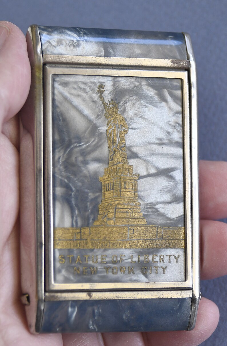 Op de afbeelding: Een rechthoekige, zilverkleurige metalen behuizing met een goudkleurig ontwerp van het Vrijheidsbeeld. De achtergrond heeft een gemarmerd, parelmoerachtig effect. De tekst "STATUE OF LIBERTY NEW YORK CITY" staat eronder.