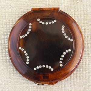 May include: A round, brown tortoiseshell compact mirror with a hinged lid. The mirror is decorated with a starburst design of clear rhinestones.