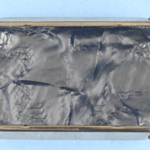 Op de afbeelding: Een rechthoekige, vintage sigarettenetui met een zilverkleurig metalen frame en een donker, gemarmerd, getextureerd oppervlak. De etui heeft een goudkleurige rand rond de randen en een kleine sluiting. Het ontwerp is eenvoudig en elegant.