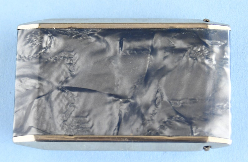 Op de afbeelding: Een rechthoekige, vintage sigarettenetui met een zilverkleurig metalen frame en een donker, gemarmerd, getextureerd oppervlak. De etui heeft een goudkleurige rand rond de randen en een kleine sluiting. Het ontwerp is eenvoudig en elegant.