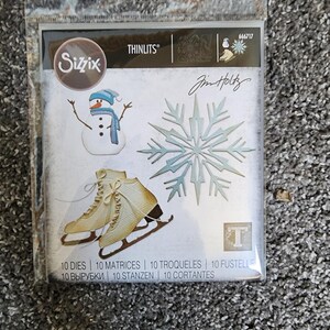 K&ouml;nnte beinhalten: Eine Packung mit 10 Metall-Stanzformen zum Basteln. Die Stanzformen zeigen einen Schneemann, eine Schneeflocke und Schlittschuhe. Die Verpackung ist mit "Sizzix Thinlits" und "Jim Holtz" beschriftet.