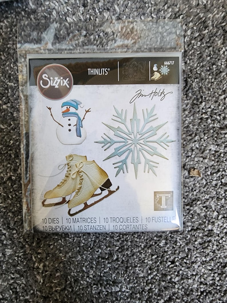 K&ouml;nnte beinhalten: Eine Packung mit 10 Metall-Stanzformen zum Basteln. Die Stanzformen zeigen einen Schneemann, eine Schneeflocke und Schlittschuhe. Die Verpackung ist mit "Sizzix Thinlits" und "Jim Holtz" beschriftet.