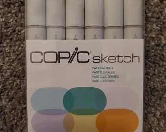 Puede incluir: Un paquete de rotuladores Copic Sketch en colores pastel. Los rotuladores tienen barriles blancos y tapas de colores. El embalaje incluye el texto "COPIC sketch" y "PALE PASTELS" en varios idiomas. La imagen también muestra el logotipo .Too.