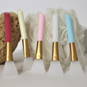 May include: Set of six silicone face masks applicators with gold handles. The applicators are in various colors: pink, light blue, white, yellow, and red. The applicators are on a white surface with a white and beige textured background.