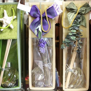 May include: Three glass diffuser bottles with reeds in decorative bags. The bags are green, beige, and beige with a purple bow. The bottles are filled with clear liquid and have a round shape. The reeds are brown and are sticking out of the bottles. The bags have different decorative elements, including a starfish, a purple bow, and a sprig of eucalyptus.