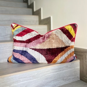 May include: A colourful geometric patterned cushion with a red border. The cushion is made of a soft, velvet-like fabric and features a variety of colours, including red, pink, blue, yellow, and orange.