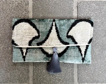 May include: A rectangular clutch with a teal and black geometric pattern. The clutch has a flap closure and a gray tassel. The pattern is a repeating design of white shapes outlined in black.