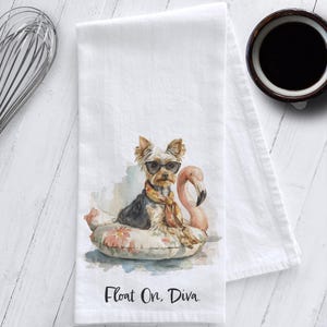 May include: White kitchen towel with a watercolor illustration of a Yorkshire Terrier wearing sunglasses and a scarf, lounging on a floral float with a pink flamingo. The text "Float On, Diva." is printed at the bottom.