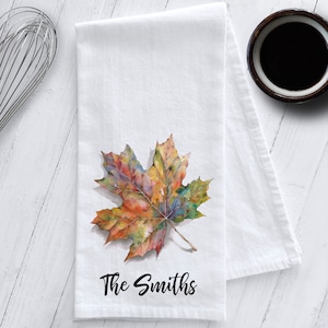 May include: White kitchen towel with a watercolor painting of a colorful fall maple leaf and the text "The Smiths" printed in black.