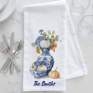 May include: White kitchen towel featuring a watercolor design of pumpkins and a blue and white vase. The vase holds a floral arrangement. The text "The Smiths" is printed in navy blue at the bottom of the towel.