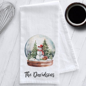 May include: White kitchen towel with a watercolor design of a snowman inside a snow globe. The snowman wears a red hat and scarf. The text "The Davidsons" is printed in black script at the bottom.