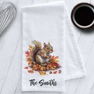 May include: A white kitchen towel with a watercolor illustration of a squirrel sitting on a pile of autumn leaves. The squirrel is holding an acorn in its paws. The text "The Smiths" is printed in black at the bottom of the towel.