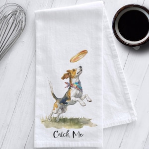 May include: White kitchen towel featuring a watercolor illustration of a beagle dog jumping to catch a frisbee. The dog is wearing a colorful bandana. The text "Catch Me" is printed at the bottom of the towel.