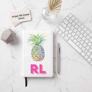 May include: A white notebook featuring a colorful watercolor pineapple illustration in shades of blue, green, yellow, and pink, with the bold pink letters RL printed below the pineapple. The notebook has a black ribbon bookmark attached at the bottom.