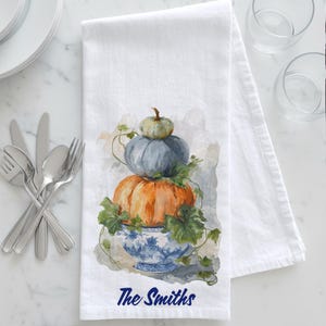 May include: White kitchen towel featuring a watercolor design of stacked pumpkins in shades of orange, blue, and green, atop a blue and white patterned bowl. The text "The Smiths" is printed in blue at the bottom.
