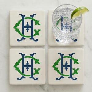 May include: Four square white ceramic coasters featuring a dark blue letter H intertwined with a green letter C. One coaster supports a clear glass filled with ice and a lime wedge, showing the monogram through the glass and liquid.