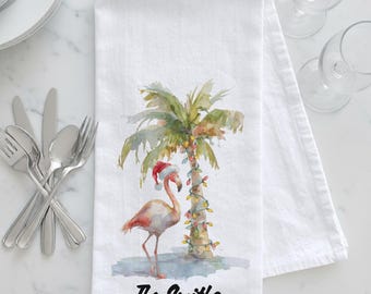 May include: White kitchen towel featuring a watercolor flamingo wearing a Santa hat, standing next to a palm tree decorated with Christmas lights. The text "The Smiths" is printed at the bottom.