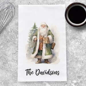 May include: A white kitchen towel with a watercolor illustration of Santa Claus holding a gift and standing next to a Christmas tree. The text "The Davidsons" is printed at the bottom of the towel.