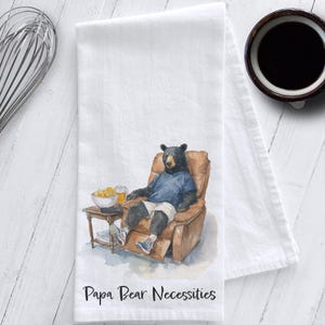 May include: White kitchen towel with a watercolor illustration of a bear relaxing in a recliner, holding a beer and snacks. The text "Papa Bear Necessities" is printed at the bottom. A whisk and a cup of coffee are in the background.