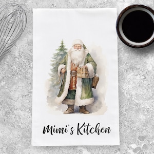 May include: A white kitchen towel with a watercolor illustration of Santa Claus holding a gift and standing next to a Christmas tree. The text "Mimi's Kitchen" is printed in black at the bottom of the towel.