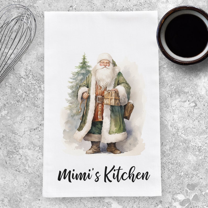 May include: A white kitchen towel with a watercolor illustration of Santa Claus holding a gift and standing next to a Christmas tree. The text "Mimi's Kitchen" is printed in black at the bottom of the towel.