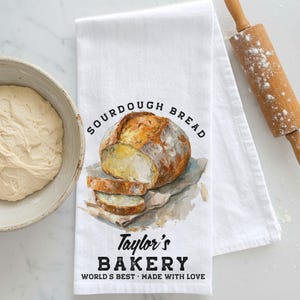 May include: White kitchen towel with a watercolor illustration of a sourdough loaf and slices. Text reads "SOURDOUGH BREAD" and "Taylor's BAKERY WORLD'S BEST MADE WITH LOVE". A bowl of dough and a rolling pin are in the background.