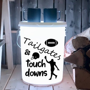May include: White plastic bucket with a black and white design that says "Tailgates & Touchdowns". The design also includes a football and a football player. Two black mugs are sitting on top of the bucket.