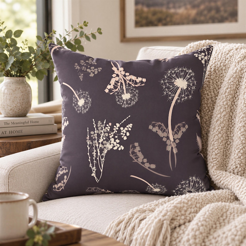 a cozy living room with a gray throw pillow adorned with white floral patterns, resting on a beige couch. The room is decorated with a vase of greenery, a stack of books, and a framed picture on the wall.