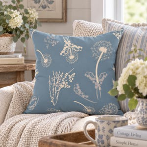 a cozy living room with a blue throw pillow adorned with white floral patterns, a wooden coffee table with a white mug and a vase of white flowers, and a bookshelf filled with books.
