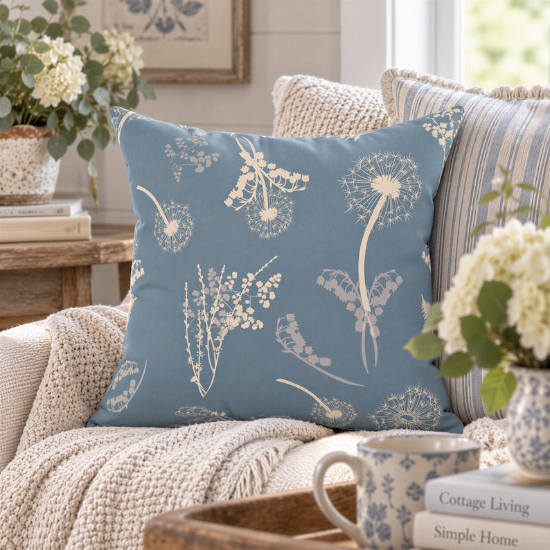 a cozy living room with a blue throw pillow adorned with white floral patterns, a wooden coffee table with a white mug and a vase of white flowers, and a bookshelf filled with books.
