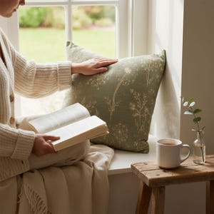 A person is sitting on a window sill, reading a book and holding a cup of tea, with a green pillow and a small vase of flowers nearby.