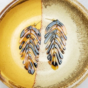Puede incluir: Un par de pendientes de carey con forma de hojas de palma. Los pendientes están en un plato de cerámica amarillo y marrón.