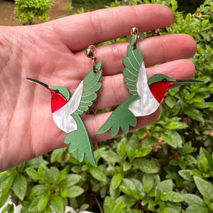 May include: A pair of green and white hummingbird earrings with red accents. The earrings are made of acrylic and have a gold-tone finish.
