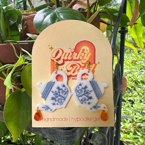 Puede incluir: Pendientes de tetera blancos y azules con un diseño floral y un dije naranja en forma de lágrima. Los pendientes se exhiben en una tarjeta color crema con el texto "Quirky Bill" y "handmade | hypoallergenic".