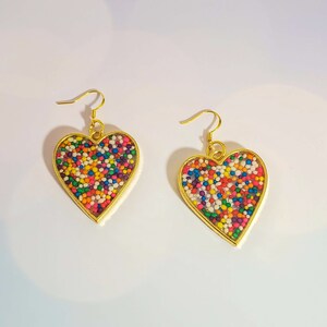 May include: A pair of gold heart-shaped earrings with colorful sprinkles embedded inside the resin.