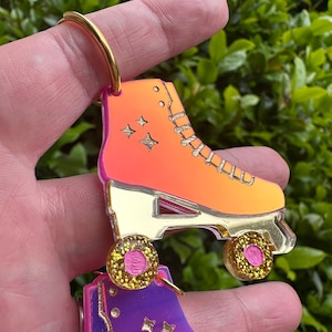 May include: A pair of roller skate keychains with gold glitter accents. The top skate is orange and pink with gold glitter wheels. The bottom skate is purple and pink with gold glitter wheels.